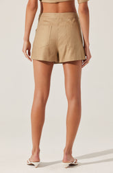 The image shows the back view of a person wearing high-waisted beige shorts made of a lightweight fabric, with two patch pockets on the back. The person is also wearing a matching beige crop top and white heeled sandals with a small block heel.